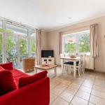 Wohnzimmer mit rotem Sofa, Essbereich und Küche. Durchgänge zu Balkon mit Gartenblick.