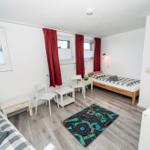 Zimmer mit zwei Betten, roten Vorhängen und Spielteppich auf Holzboden.