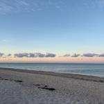 Strand mit feinem Sand und ruhigem Meer unter einem klaren Himmel mit rosa gefärbten Wolken.