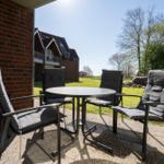 Terrasse mit Tisch und vier Stühlen. Hintergrund: Grasfläche, Bäume und Gebäude.