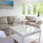 Wohnzimmer mit Sofa, Sessel und Glas-Tisch. Große Fenster und Wandbild mit Strandmotiv.