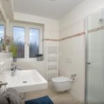 Badezimmer mit Dusche, Waschbecken und Toilette. Fenster und Handtuchhalter sichtbar.