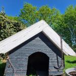 Holzhütte mit Schieferdach im Garten, umgeben von Bäumen und Pflanzen.