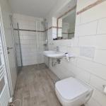 Bathroom with shower, sink, and toilet. White tiles and wooden floor.