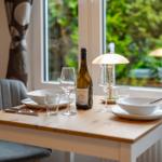 Esstisch mit Geschirr, Weinflasche und Lampe vor Fenster mit Blick auf Garten.