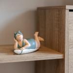 Eine kleine Figur liegt auf einem Brett auf einer Holzregalstufe.