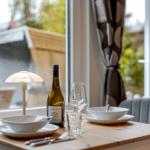 Esstisch mit Geschirr, Wein und Lampen vor Fenster mit Gartenblick.