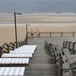 Holzterrasse mit Liegestühlen führt zum Strand. Zwei Personen stehen am Geländer.