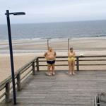 Zwei Figuren stehen auf einer Holzterrasse mit Blick auf den Strand und das Meer.
