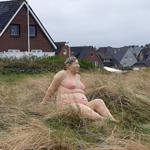Eine Skulptur einer Frau im Bikini sitzt im hohen Gras. Hintergrund: Wohnhäuser und bewölkte Himmel.