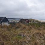 Dorfhaus mit Strand im Hintergrund, umgeben von Dünen und hohem Gras.