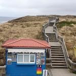Blauer Strandkiosk mit rotem Dach und Holzsteg zur Dünenlandschaft.
