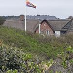 Haus mit Schindeldach und Flagge auf Mast. Umgeben von Grünflächen.