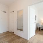 Flur mit Spiegel und Türen. Durch eine Tür ist ein Schlafzimmer mit Stuhl und Nachttisch sichtbar.