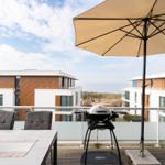 Terrasse mit Tisch, Stühlen, Sonnenschirm und Grill. Blick auf moderne Gebäude und Strand.