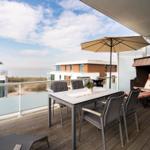 Terrasse mit Tisch, Stühlen, Sonnenschirm und Liege. Blick auf Wohngebäude und Meer.