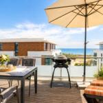 Terrasse mit Tisch, Stühlen, Grill und Sonnenschirm. Blick auf Meer und moderne Gebäude.