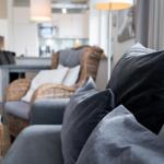 Ein Wohnbereich mit grauem Sofa und Kissen, einem Stuhl aus Rattan und einer Küchenzeile im Hintergrund.