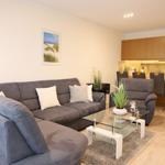 Wohnbereich mit grauem Sofa, Glas-Tisch und Küchenbereich im Hintergrund.