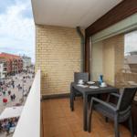 Balkon mit Tisch und Stühlen, Blick auf belebte Straße mit Geschäften und Passanten.