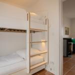 Bunk bed with white mattresses and bedside lamps. Living room with sofa in background.