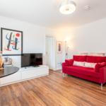 A living room with red sofa, wooden floor, and TV on white cabinet.