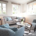 Wohnzimmer mit Sofa, zwei Sesseln und runden Tischen. Drei Fenster mit Blick auf Grün.