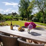 Holztisch mit Stühlen, Blumen und Obst auf Terrasse mit Gartenblick.