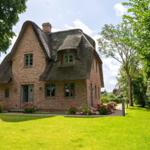 Haus mit strohgedecktem Dach und grünem Garten mit Blumenbeeten.
