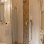 Dusche mit Glaswand, Duschbrause und Handbrause. Spiegel, Toilette und Heizkörper sichtbar.