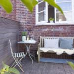 Terrasse mit Holzbank, Kissen und Tisch vor Backsteinmauer.