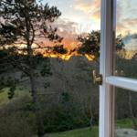 Durch das Fenster blickt man auf einen Garten mit Bäumen und einen Sonnenuntergang.