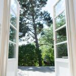Durch offene Fenster blickt man auf eine Terrasse mit Hängesessel und einen hohen Baum.