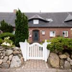 Red brick building with white door and garden gate. Surrounded by shrubs and stones.