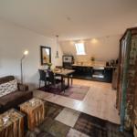 Living and dining area with kitchen under sloped ceiling. Wooden floor, sofa, table, and cabinet visible.