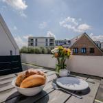 Terrasse mit Tisch, Stühlen, Brot und Blumen. Blick auf Wohnhäuser und Himmel.