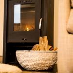 Holzofen mit brennendem Feuer und Korb mit Brennholz vor einem Sofa.