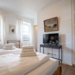 Two beds with white sheets, windows with white curtains, and TV on table.