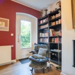 Room with red wall, bookcase, armchair, and fireplace.