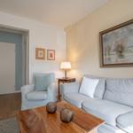 Living room with sofa, armchair, and wooden table. Wall painting and lamp visible.