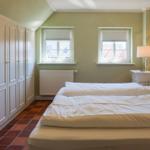 Schlafzimmer mit Bett, Kleiderschrank und zwei Fenstern.