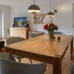 Dining room with wooden table, chairs, and flower arrangement. Windows and door lead outside.