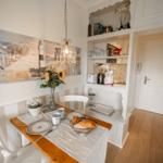 Dining table with tablecloth, mugs, and bread. Kitchenette and wall art with pendant light.