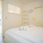 Schlafzimmer mit Bett, Handtuch und Tür. Durchgang zu Wohnbereich mit Tisch und Stuhl.