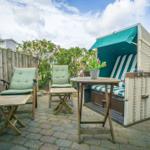 Terrasse mit Strandkorb, Tisch und zwei Stühlen. Holzplatten und grüne Kissen.