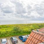 Vom Balkon aus Blick auf den Strand und das Meer mit grünen Dünen und geparkten Autos.