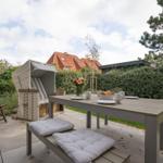 Terrasse mit Tisch, Stühlen und Strandkorb. Hinterhof mit Hecke und Bäumen.
