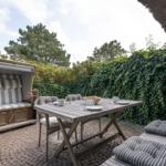 Terrasse mit Holztisch, Stühlen und Strandkorb. Hintergrund: grüne Hecke und Bäume.