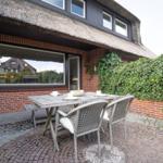Terrasse mit Tisch und Stühlen vor einem Haus mit Schilfdecke und Kletterpflanzen.