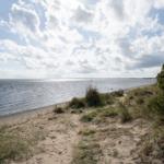 Strand mit Dünen, Gras und Steinmauer unter bewölktem Himmel.